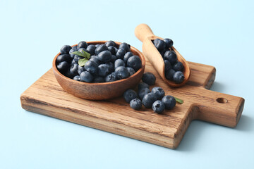 Wooden cutting board, scoop and bowl with fresh ripe blueberry on blue background, closeup