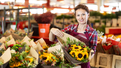 Positive middle-aged saleswoman holding bouquet of sunflowers in flower market
