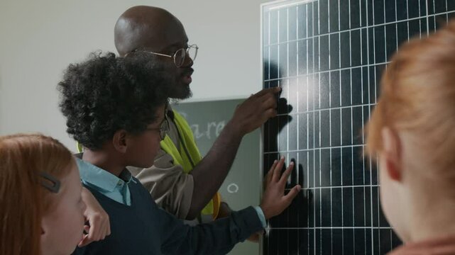 Medium close-up of black male engineer and group of diverse children studying solar panel at school career day, touching its surface, discussing its configuration, functioning and work principles