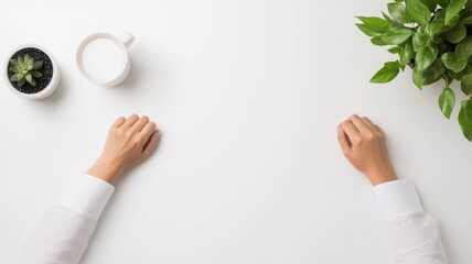 Obraz premium An aerial view of hands resting on a white desk with a plant, cup, and minimalistic workspace for a serene atmosphere.