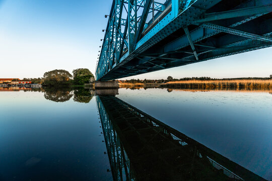 Underneath the blue bridge towering over the biggest river in Randers, Denmark near wildlife marsh at between blue hour and golden hour with still water reflection of bridge POV