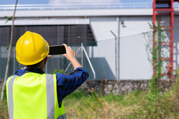 An engineer in a hard hat and safety vest conducts a site inspection, using a smartphone to photograph an industrial building for a report.