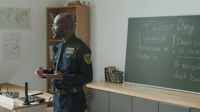 Medium shot of confident black policeman in blue uniform giving lecture about personal safety to schoolchildren at visit on career day, standing in front of blackboard with rules handwritten in chalk