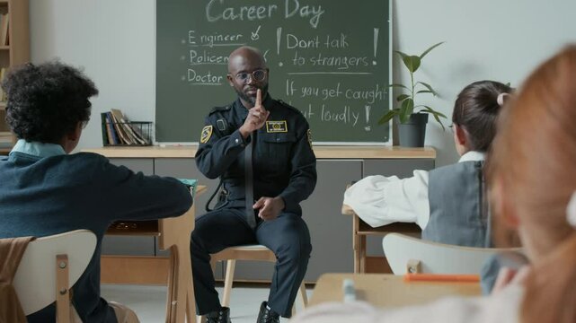 Medium shot of young African American police officer in uniform sitting on chair in classroom with 10-year-old diverse students at school, chatting about safety precautions and dealing with criminals
