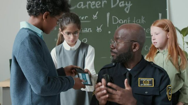 Medium shot of young African American police officer in uniform sitting in classroom, surrounded by multiethnic 10-year-old kids, telling about profession, answering questions, giving badge to look at