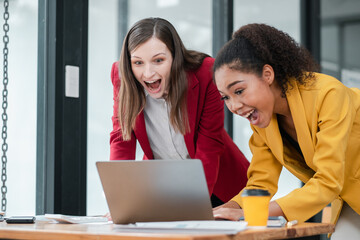 Two enthusiastic businesswomen in professional attire working together on a laptop in a contemporary office setting.