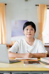 A young student sitting at a desk with a laptop, books, and a notebook, studying in a bright room with yellow curtains.