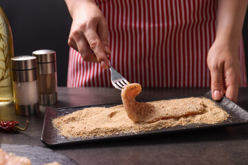 Making schnitzel. Woman coating slice of meat with bread crumbs at dark table, closeup