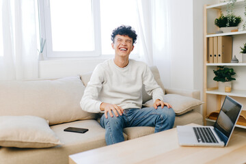 Young man smiling while sitting on a couch in a bright, airy room with a laptop nearby Natural light enhances the cozy atmosphere with indoor plants