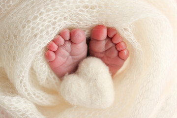 The tiny foot of a newborn baby. Soft feet of a new born in a white wool blanket. Close up of toes, heels and feet of a newborn. Knitted white heart in the legs of a baby. Macro photography. 