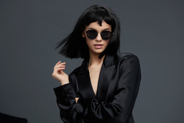 Woman posing in black suit and sunglasses on gray background, with hand on hip and head tilted,...