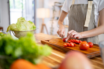 A cropped image of a woman slicing a tomato on a wooden cutting board at the kitchen table.