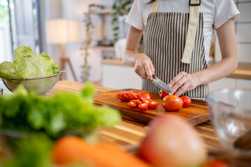 A cropped image of a woman slicing a tomato on a wooden cutting board at the kitchen table.