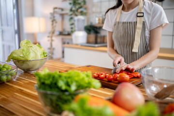 A cropped image of a woman slicing a tomato on a wooden cutting board at the kitchen table.