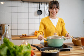 A beautiful Asian woman is cooking in the kitchen, placing a pot on an electric portable stove.