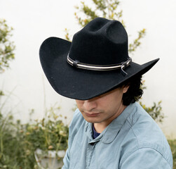 Mestizo male model with curly hair wearing blue shirt and wool cowboy hat with leather decoration. Portrait with blurred natural background and contemporary Andean cultural style.