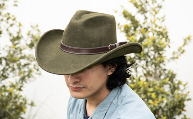 Mestizo male model with curly hair wearing blue shirt and wool cowboy hat with leather decoration. Portrait with blurred natural background and contemporary Andean cultural style.