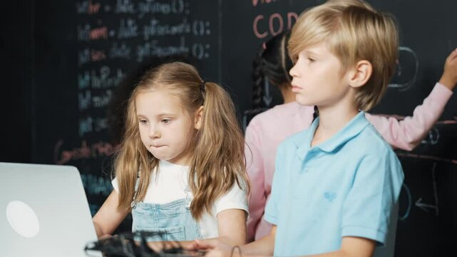 Smart girl using laptop programing engineering code with diverse friends ay table with microchip and main board scatter around in STEM classroom at blackboard written with coding prompt. Erudition.