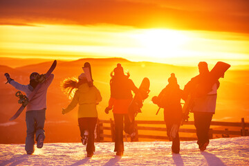 Fototapeta premium Group of happy young friends are having fun and runs with ski and snowboards in hands at sunset mountain top. Ski resort and winter vacations concept