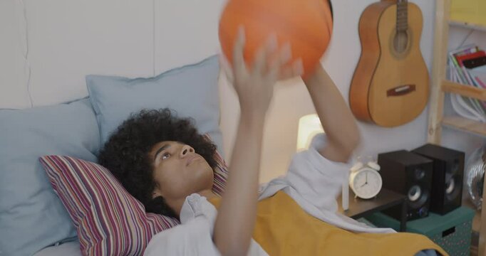 Bored African American teenager lying in bed throwing and catching basketball spending time at home. Leisure activity and teenage lifestyle concept.
