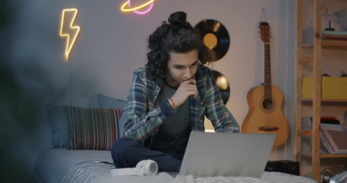 Happy biracial teenager using laptop computer typing sitting in bed in bedroom in evening. Modern technology and adolescence concept.