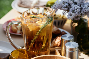Refreshing iced tea served in clear pitcher adorned with lemon slices and decorative rosemary next to flowers and seasoning containers on outdoor table setup