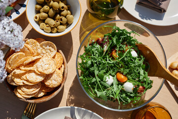 Display of Mediterranean salad with arugula, cherry tomatoes, mozzarella balls, and olives in glass bowl, accompanied by crackers and other side dishes on outdoor dining table setting