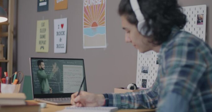 Teenage boy studying in online school watching video lesson in virtual classroom and writing in notebook at home. Distance education and modern technology concept.