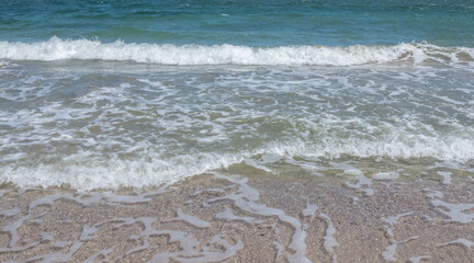 Beautiful summer seascape. Blue sea water surface with green and turquoise tones and shades, foamy waves on the coast, wet beige sand on the beach. Natural colorful pattern, suitable for backgrounds.