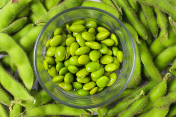 A glass bowl is filled with vibrant green edamame beans, with additional beans scattered around on a white background