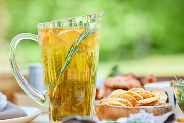 Refreshing citrus-infused beverage in elegant glass pitcher with rosemary sprig, accompanied by snacks on wooden table in bright outdoor setting with blurred background