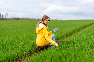 A young farmer woman in a  green field, intently using a tablet. She wears a yellow jacket and glasses, enjoying the serene, cloudy day outdoors. The concept of technology, gardening.