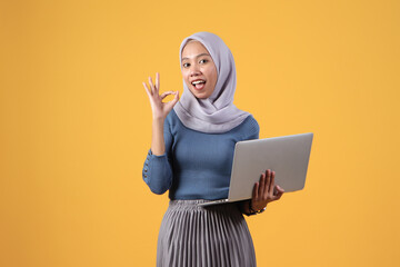 happy asian indonesian muslim woman holding laptop computer giving ok finger sign on isolated yellow background