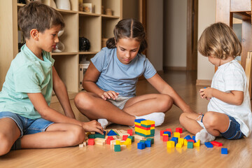 Obraz premium Family Time, Early Childhood Education, And Fun At Home. Happy Smiling Preschool Or Elementary School Age Siblings Or Friends Sitting On The Floor Playing And Building With Wooden Blocks.