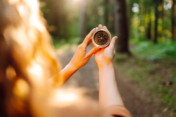 A hiker with curly hair holds a compass in their hands, carefully navigating through a sunlit forest filled with tall trees, greenery, and warm afternoon light. © maxbelchenko