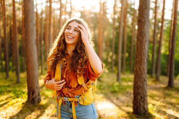 Happy young woman with curly hair walks along a forest path, wearing a yellow hiking backpack surrounded by tall trees and warm sunlight. Trips. Active lifestyle. Hiking.