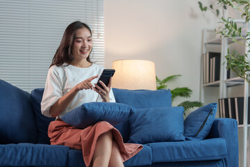 Happy and relaxed Asian woman sitting on sofa on weekend using smartphone to communicate, talk, text, chat online, watch wireless internet on device. Shopping online from your home office.