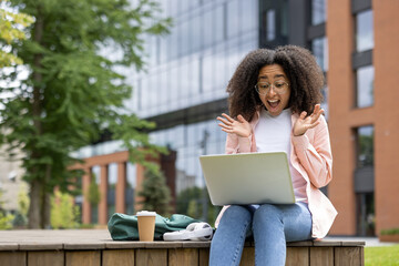 Joyful woman sitting outdoors using laptop, expressing excitement and surprise. Urban background with glass buildings, casual clothing, enjoying coffee. Scene captures remote work, technology