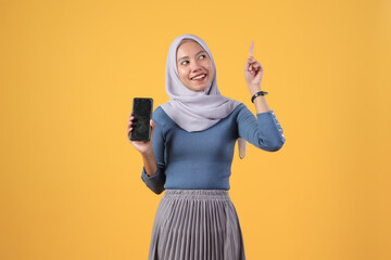 happy asian indonesian muslim woman holding smart phone showing blank screen and giving pointing finger gesture on isolated yellow background