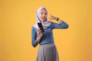 happy asian indonesian muslim woman holding smart phone showing blank screen and giving thumb up gesture on isolated yellow background