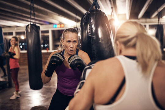 Two focused mature women sparring with gloves in a boxing gym. There are punching bags hanging from the ceiling behind them.