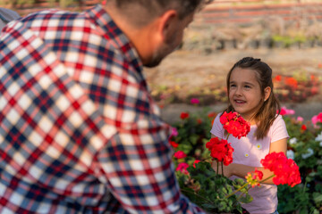 A father handing a flower pot to his smiling daughter in a greenhouse.