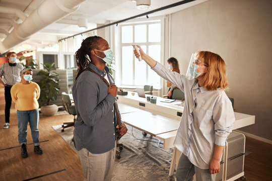 Black businessman having his temperature read, by a colleague in an office community before he is allowed to enter the workplace. His colleagues are queuing up behind him