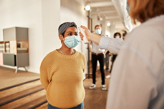 Young female entrepreneur in a yellow shirt, having her temperature taken, by a colleague in an office community before she is allowed to enter the workplace. Her colleagues are queuing up behind her