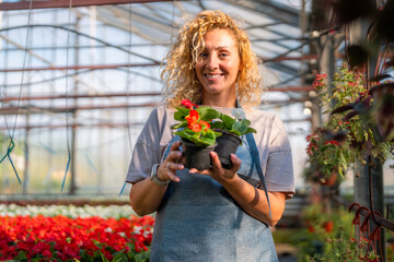 Woman with curly hair proudly holding a potted plant in a well-maintained greenhouse.