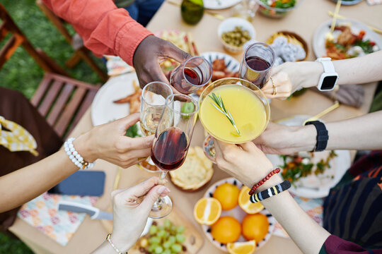 Group of friends raising glasses for toast during outdoor gathering with diverse food on table, showcasing various cuisines, drinks and lively atmosphere on sunny day
