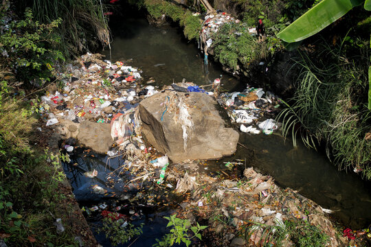 A close-up photo of a river polluted with various types of trash, including plastic bags, bottles, and food wrappers. The trash is piled up on the riverbanks and in the water, which is dark and murky