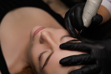 Young beautiful girl lying relaxed at the procedure of permanent eyebrow makeup, the master applies...