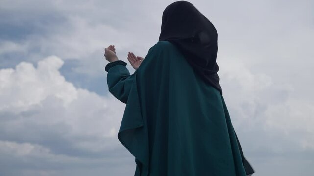 Muslim Woman Wearing a Dress and Hijab Prays Devoutly with Hands Raised Outdoors Under the Sky. Concept of Islam, faith, spirituality, hope, worship, gratitude, dua, mindfulness