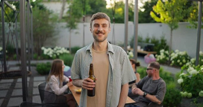 Portrait of a happy blond guy with a mustache with a bottle of brown beer in his hands while relaxing with a company in the backyard of a country house